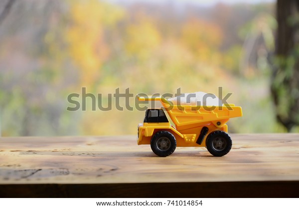 A small yellow toy\
truck is loaded with a stone of white salt. A car on a wooden\
surface against a background of autumn forest. Extraction and\
transportation of salt