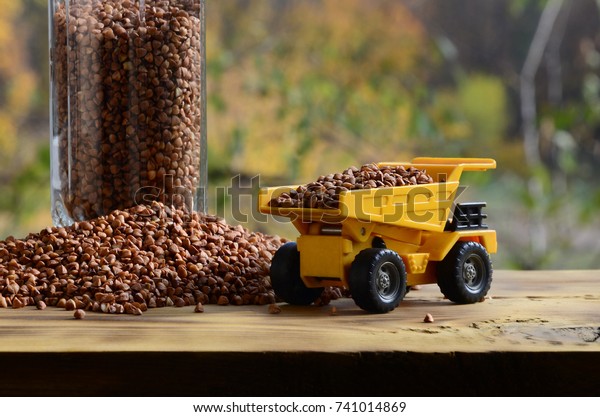 A small yellow toy truck is loaded with brown\
grains of buckwheat around the buckwheat pile and a glass of croup.\
A car on a wooden surface against a background of autumn forest.\
Buckwheat delivery