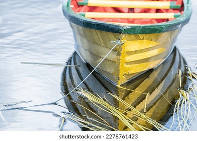 A small yellow wooden row boat with green trim and a red interior floats on calm water with reeds in the water. The boat is tied on with green rope. The boat's reflections are seen in the water.