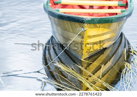 A small yellow wooden row boat with green trim and a red interior floats on calm water with reeds in the water. The boat is tied on with green rope. The boat's reflections are seen in the water.