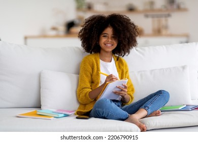 Smart African American Girl With Bushy Hair Schooler Sitting On Couch, Holding Notepad And Pen, Writing Essay And Smiling, Kid Doing Homework, Enjoying Educational Process, Home Interior, Copy Space