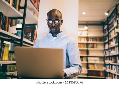 Smart African American guy in round glasses typing on laptop keyboard while sitting near bookcase and doing homework in library