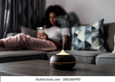 Smart air humidifier emitting water vapor on table near sofa with black female at home