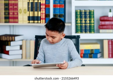 Smart Asian boy on grey sweatshirt sit at school desk, lower head down to concentrate on online class study of kid lesson by reading ebook, info searching via tablet at library of elementary academy