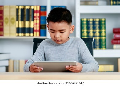 Smart Asian boy on grey sweatshirt sit at school desk, lower head down to concentrate on online class study of kid lesson by reading ebook, info searching via tablet at library of elementary academy