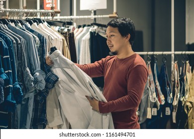 Smart Asian Man With Beard Choosing Clothes In Clothing Store At Shopping Center, Looking White Color T-shirts , Fashion And Consumerism Concept.