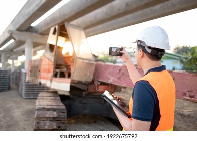 Smart Asian worker man or male civil engineer with protective safety helmet and reflective vest holding digital tablet using smartphone for taking photo of excavator machine at construction site.