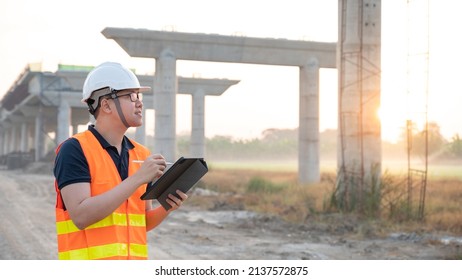 Smart Asian Worker Man Or Male Civil Engineer With Protective Safety Helmet And Reflective Vest Using Digital Tablet For Project Planning And Checking Architectural Drawing At Construction Site.