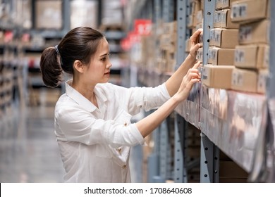 Smart Asian Young Woman Happy To Work In Store Warehouse. Girl Standing And Checking Quality And Quantity Of Inventory Stock On Shelf By Model Name And Serial Number It Show On Packaging Of Goods. 