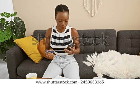Smart, beautiful african american woman sitting on sofa seriously concentrating on phone, typing a text message for online shopping at home, using credit card for payment