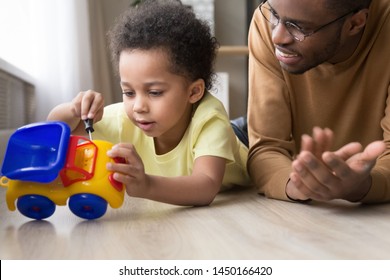 Smart Biracial Preschooler Boy Lying On Floor With Young Black Dad Repair Toy Car With Plastic Wrench, Loving African American Father Play With Little Child At Home Teach Fixing Maintenance Vehicle