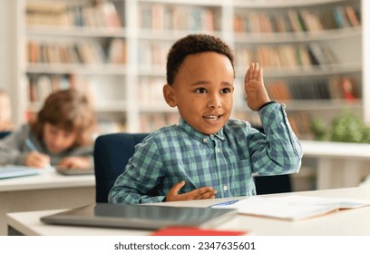 Smart black boy raising his hand to answer a question during lesson in elementary school classroom, kid engaging in the lesson led by his educator - Powered by Shutterstock