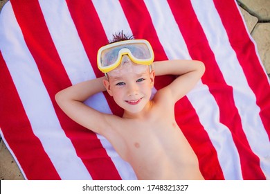 Smart blue-eyed little boy in yellow diving goggles lying on Striped red and white beach towel and smiling happily. Copy space