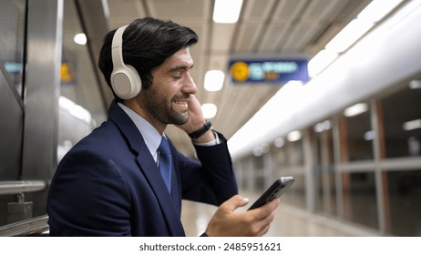 Smart business man listening music by headphone while waiting train at train station with blurring background. Skilled project manager enjoy listen relax sound while holding mobile phone. Exultant. - Powered by Shutterstock