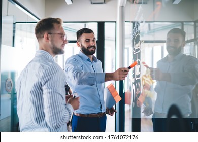 Smart casual businessmen discussing organisation plan from board during working time in company, happy Caucasian male employees creating productive strategy while analyzing information indoors