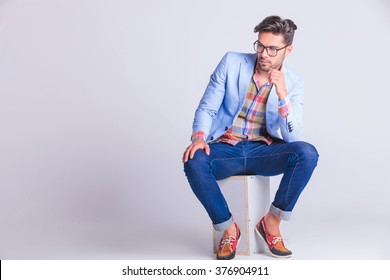 Smart Casual Man Seated On Box, Wearing Glasses And Jeans, Posing While Looking Away In Studio Background