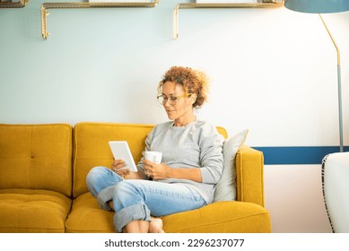 Smart casual woman middle age relaxing at home sitting on yellow sofa and using ereader to read an ebook enjoying indoor leisure activity alone. Sunday time lifestyle. People and technology online