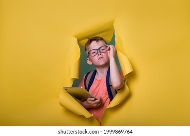 Smart Child In Glasses Holding Laptop Computer Tablet And Breaking Through Yellow Paper Wall. Back To School. Student With Backpack. Nerd Kid Portrait. Programming IT And Developer Lessons.