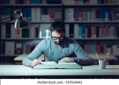 Smart confident young man studying late at night, he is sitting at desk and reading a book, knowledge and learning concept - Powered by Shutterstock