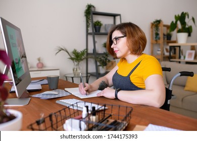 Smart disabled girl in wheelchair sitting at desk and making notes in workbook while listening to online university course at home