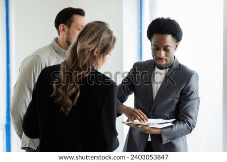 Smart ellegant african american dark skinned real estate in new empty building holding documents of the building showing house to young family couple future ownerships choosing home for family. Stock photo © 
