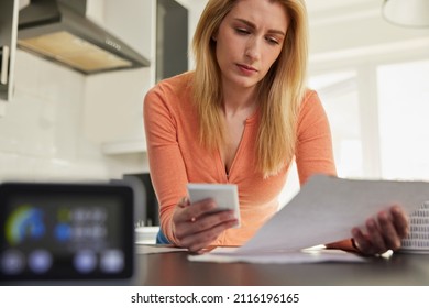 Smart Energy Meter In Kitchen Measuring Electricity And Gas Use With Woman Looking At Bills With Calculator