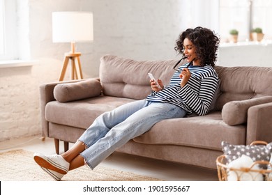 Smart ethnic woman biting glasses and browsing smartphone while resting on comfortable couch at home - Powered by Shutterstock