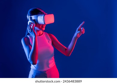 Smart female stand with surrounded by cyberpunk neon light wear VR headset connecting metaverse, futuristic cyberspace community technology. Woman using finger pointing virtual object. Hallucination. - Powered by Shutterstock