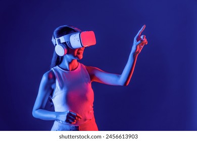 Smart female stand with surrounded by cyberpunk neon light wear VR headset connecting metaverse, futuristic cyberspace community technology. Woman using finger pointing virtual object. Hallucination. - Powered by Shutterstock