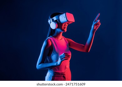 Smart female stand with surrounded by cyberpunk neon light wear VR headset connecting metaverse, futuristic cyberspace community technology. Woman using finger pointing virtual object. Hallucination. - Powered by Shutterstock