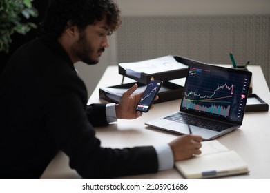 Smart Focused Indian Broker Investor, Stock Trader Using Smartphone And Laptop To Analyze And Invert In The Financial Market Of Cryptocurrencies, Buy Or Sell Crypto Coins, Planning Strategy