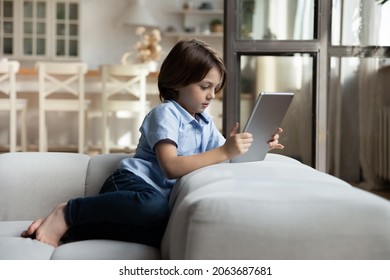 Smart Focused Primary School Boy Reading Book On Tablet Computer On Sofa. Kid Using Online App On Digital Device Alone, Watching Video Content, Playing Video Game At Home. Addiction In Childhood