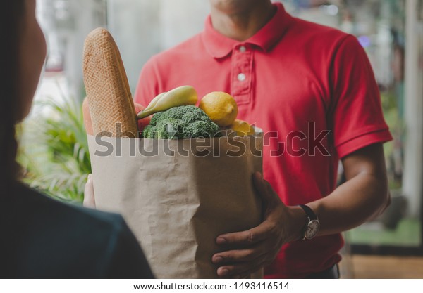smart food delivery service man in red uniform handing\
fresh food to recipient and young woman customer receiving order\
from courier at home, express delivery, food delivery, online\
shopping concept 