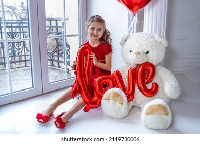 a smart girl in a red dress with red gel balloons and a white large toy teddy bear near a large arched window. Beautiful child congratulates on the holiday