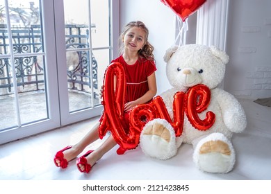 a smart girl in a red dress with red gel balloons and a large white toy teddy bear near a large arched window in a festive mood congratulates on Valentine's Day and March 8