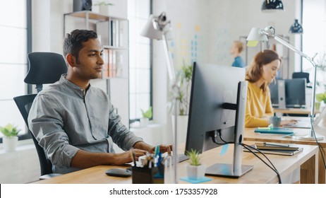 Smart and Handsome Indian Office Worker Sitting at His Desk works on a Laptop. In the Background Modern Office with Diverse Team of Young Professionals Working.