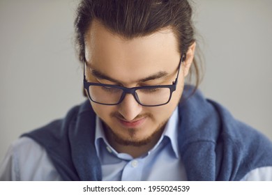 Smart Handsome Young Guy Being Shy. Closeup Portrait Of A Man In Glasses And An Office Shirt With A Ponytail Looking Down On A Gray Studio Background