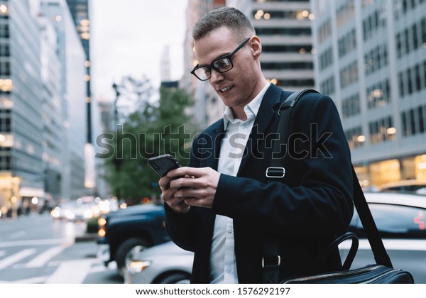 Smart and happy entrepreneur in formal suit\
and glasses walking around city and messaging in social media on\
blurred background