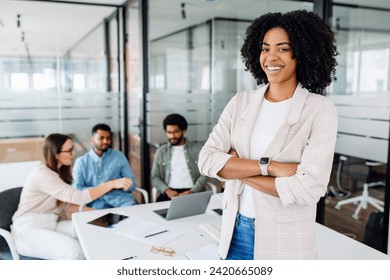 Smart high-skilled young Brazilian woman with a cheerful smile stands with arms crossed on the forefront in a modern office setting with her colleagues collaborating in the background, team synergy - Powered by Shutterstock