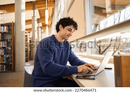 Smart Hispanic male student sitting at desk and using laptop, preparing diploma project in library campus. People. Education. Modern wireless technology. E-learning and distance communication concept