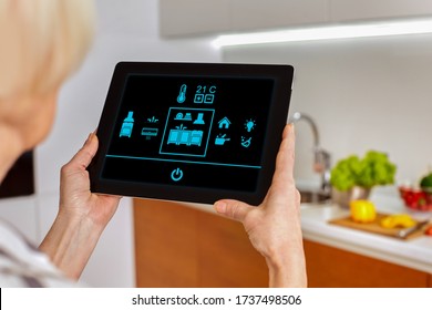 Smart home system kitchen control! Picture from back blurred background old woman hold tablet look on icons monitor device in fonteground. Touch fingers manage temperature room house interior. Mockup - Powered by Shutterstock