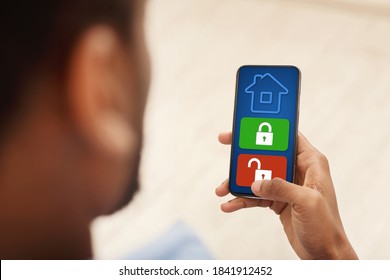 Smart House, Home Automation, Device With App Icons. Over The Shoulder View Of Black Man Using His Smartphone With Home Security Application On The Screen To Unlock The Door Of The House