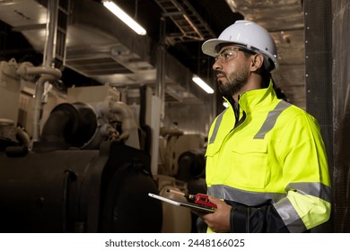 Smart HVAC system eingeer with green safety jacket and PPE hold digital tablet and and walkie talkie working at site line of HVAC control room 