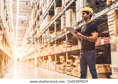 Smart Indian engineer man worker wearing safety helmet doing stocktaking of product management in cardboard box on shelves in warehouse. Factory physical inventory count.