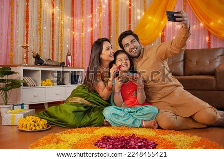 Smart Indian family taking a selfie with smartphone near Diwali rangoli: Festival and technology concept. Sweet little girl holding a tray of flowers sitting with mother and father posing for a sel...