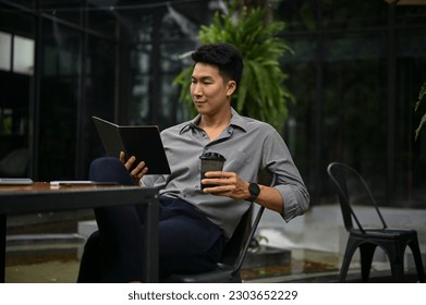 Smart and inspired millennial Asian businessman sits outdoors, enjoying his coffee, and reading a book in a coffee shop.