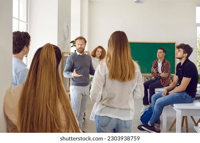 Smart and intelligent male teacher speaks in classroom surrounded by his high school students. Man talks to students at break or informal meeting where they discuss ideas about educational process. - Powered by Shutterstock