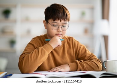 Smart korean teen kid chubby boy with glasses doing homework at home, sitting at table with books and notebooks, holding pen and touching his chin, doing exercises, copy space
