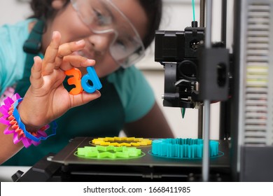 A smart latino student wearing multi-colored 3d printed shapes as jewelry, next to a 3d printer with designs on the heated print bed, and holding 3d letters in her hand.