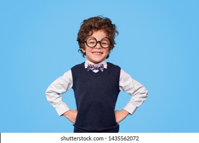 Smart little boy with curly hair wearing nerdy glasses and school uniform smiling for camera while keeping hands on waist against blue background - Shutterstock ID 1435562072